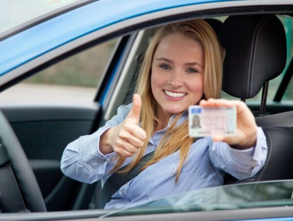 A woman in the drivers seat of her car holding up her license.